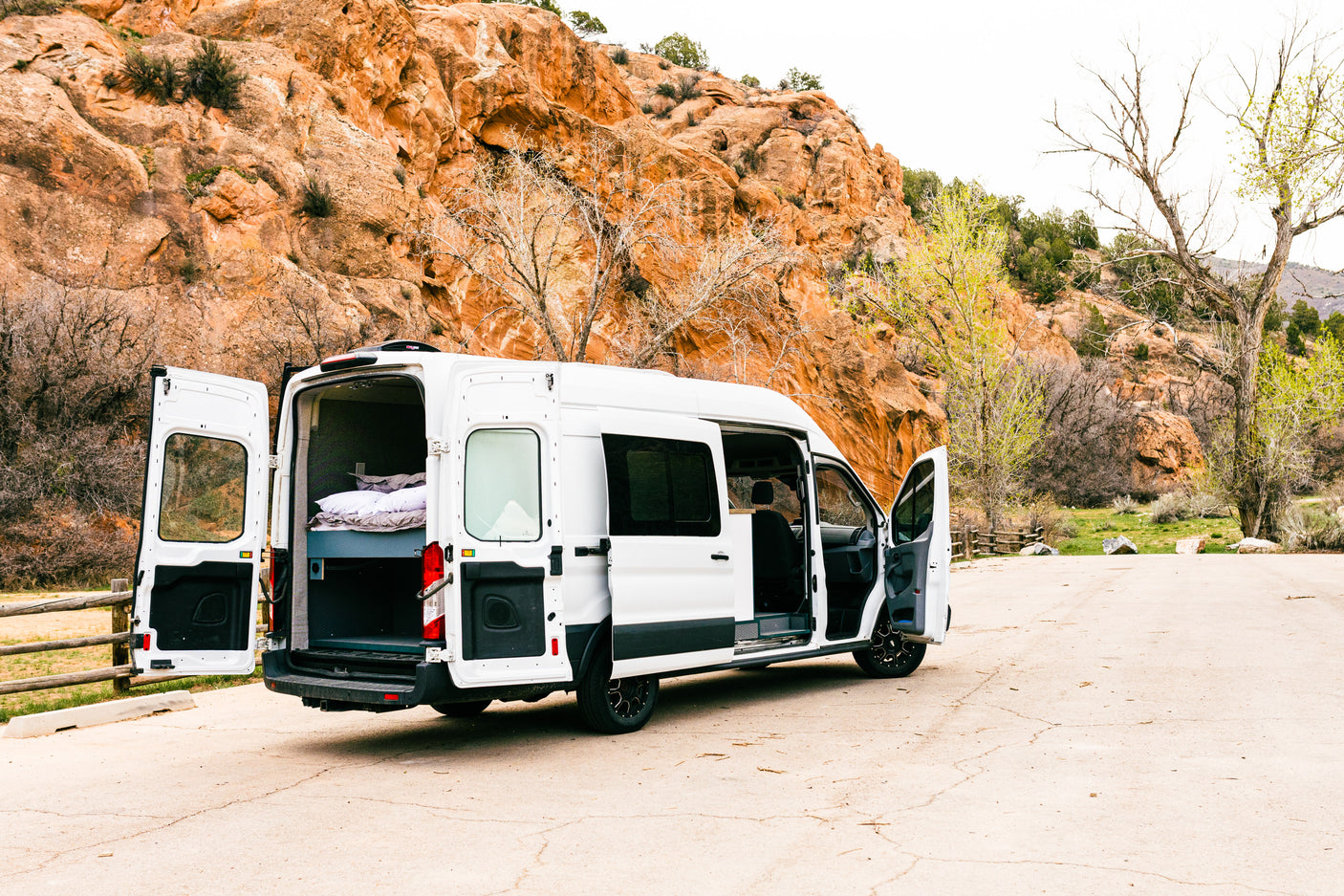White camper van (RV rental) parked by red rock cliffs. Open doors reveal a cozy bed inside, with trees surrounding.