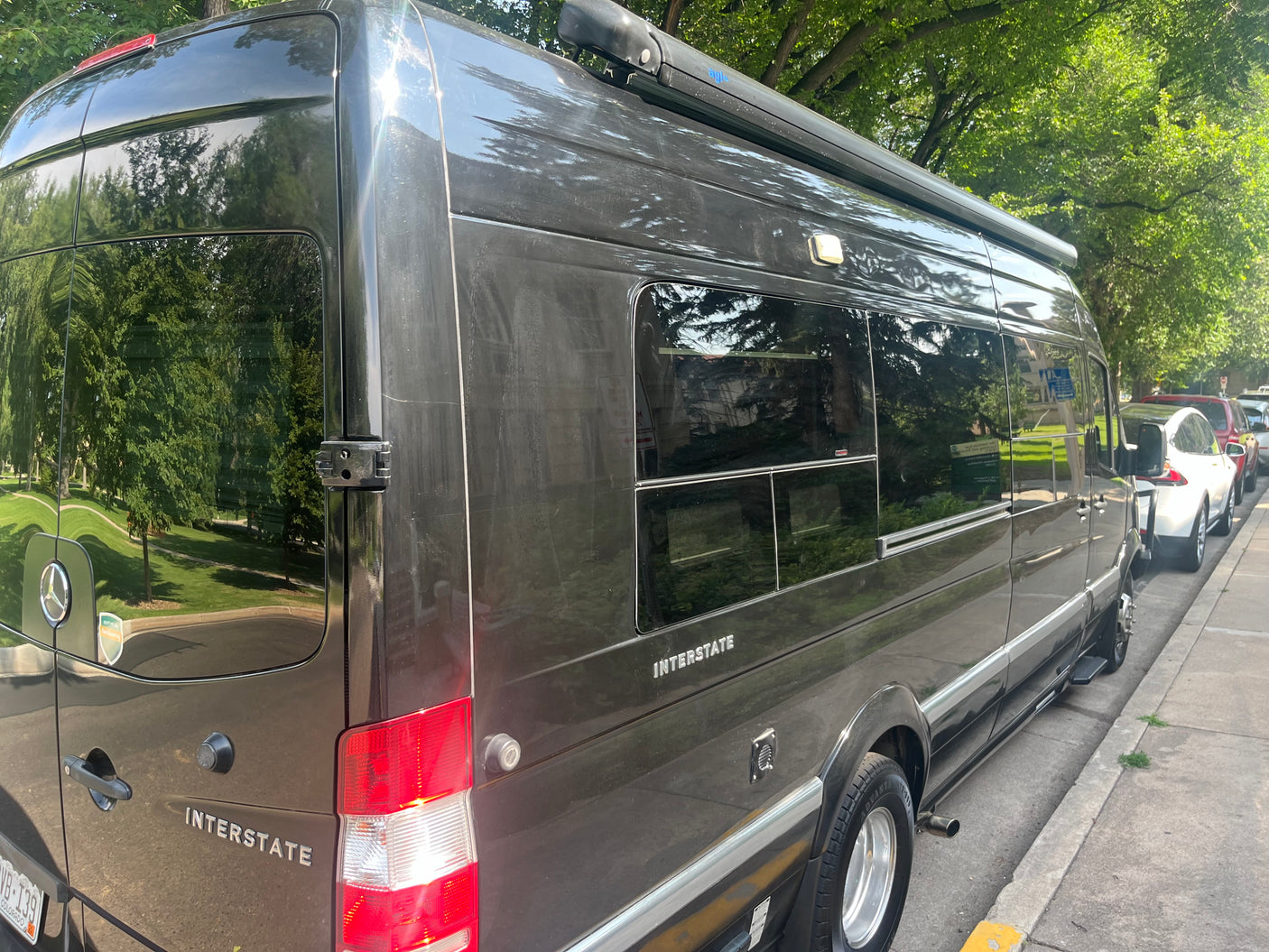 This dark grey Mercedes-Benz Interstate RV rental van is parked curbside, reflecting green trees.