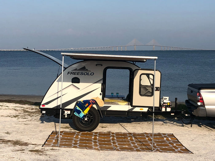 A Freesolo teardrop camper (RV rental) with awning extended on a sandy beach, overlooking a long bridge and calm water.