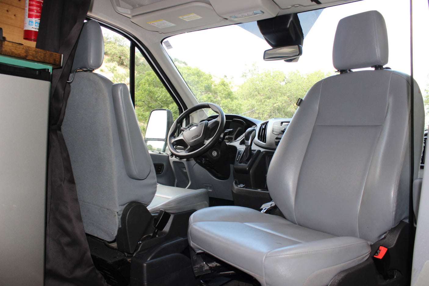 Interior of an rv rental van, showing driver and passenger seats, steering wheel, and dashboard with a view of trees outside.