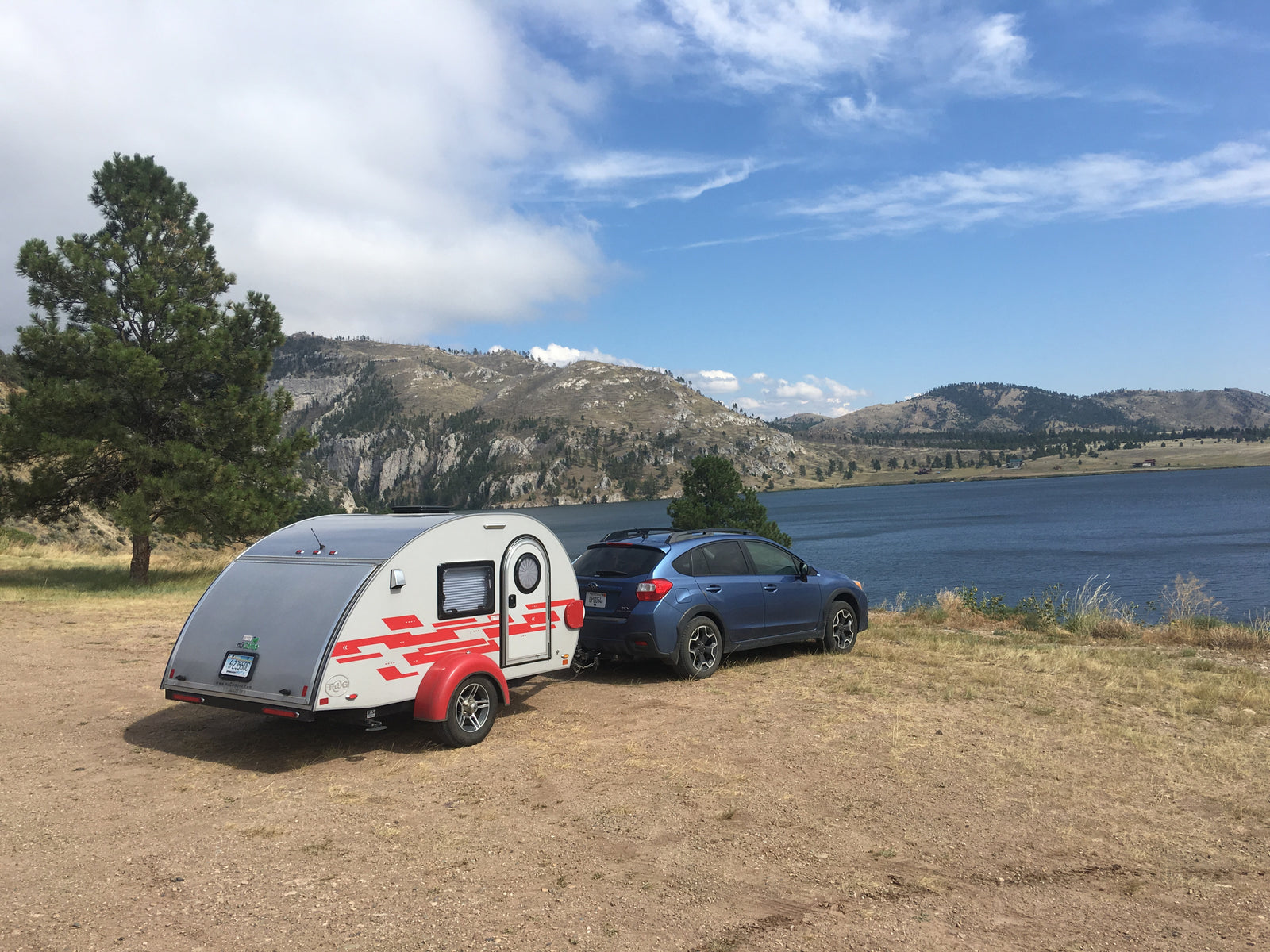 A blue Subaru tows a grey teardrop RV rental, parked by a scenic lake and mountains for camping/travel.