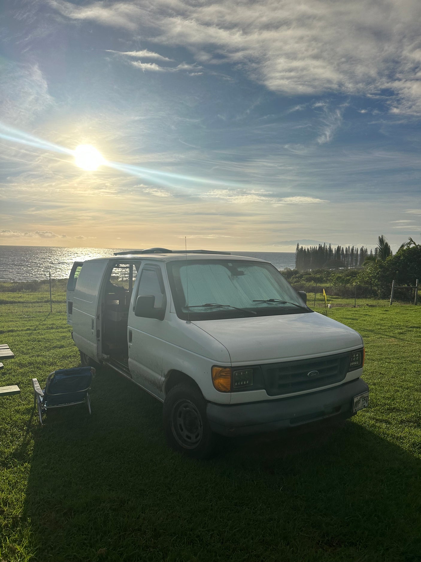 White camper RV rental van parked on green grass by the ocean at sunset, side door open for a scenic travel experience.
