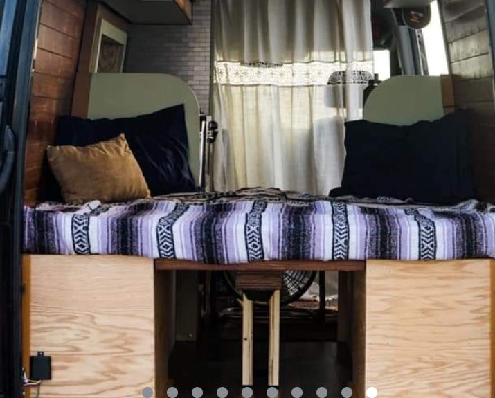 RV rental van interior: A cozy bed with a striped blanket & pillows, flanked by wood walls. Under-bed storage visible. White curtains in back.