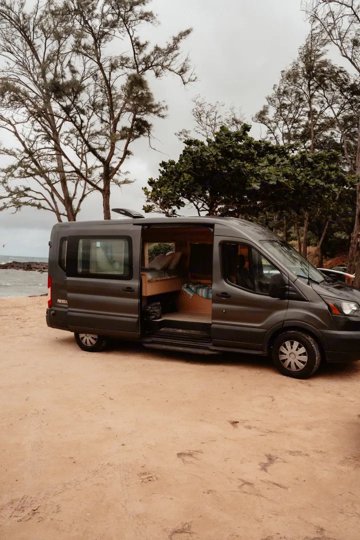 RV rental: A dark grey camper van parked on a sandy beach. Its open door reveals a cozy wooden interior, perfect for coastal travel.