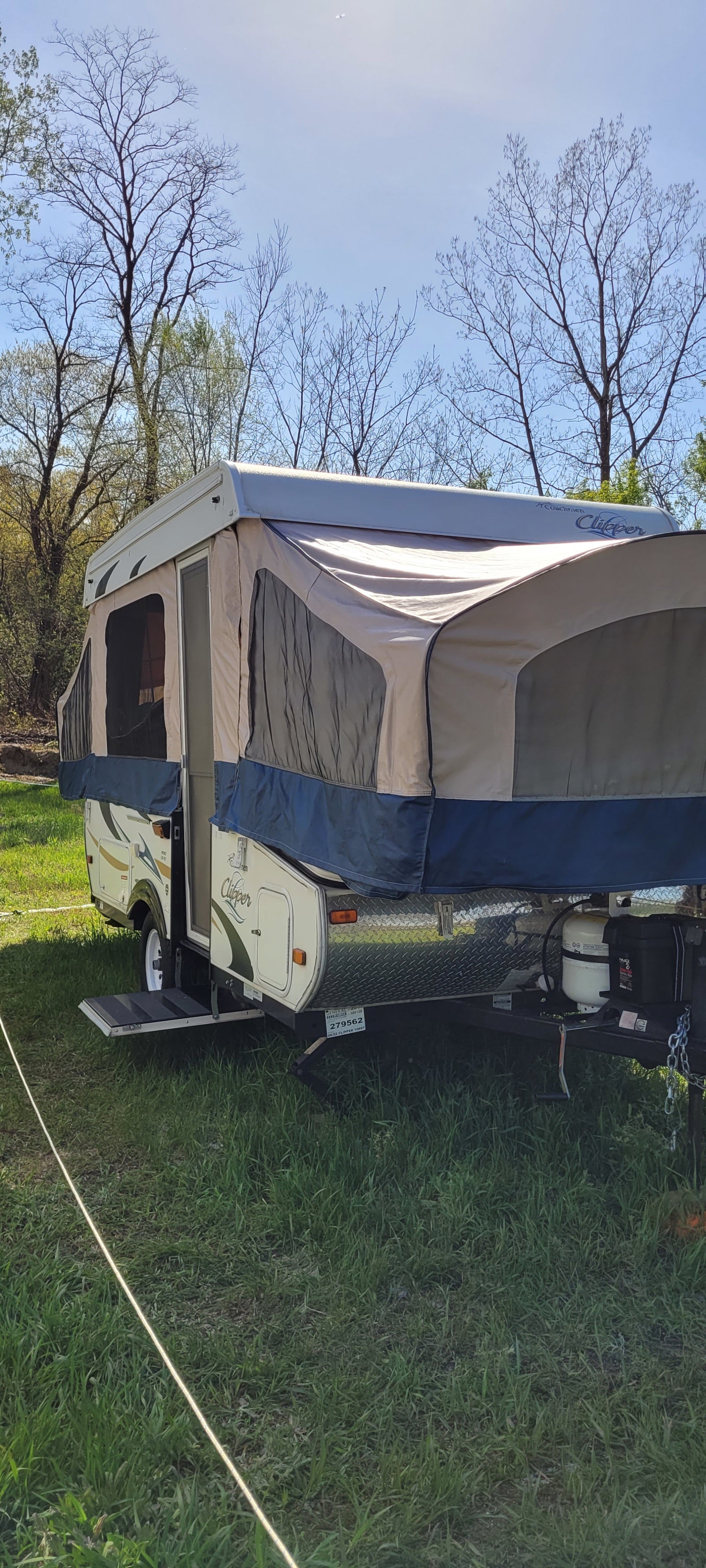 A cream and blue pop-up camper RV rental is set up in a sunny, grassy campsite with bare trees against a blue sky.