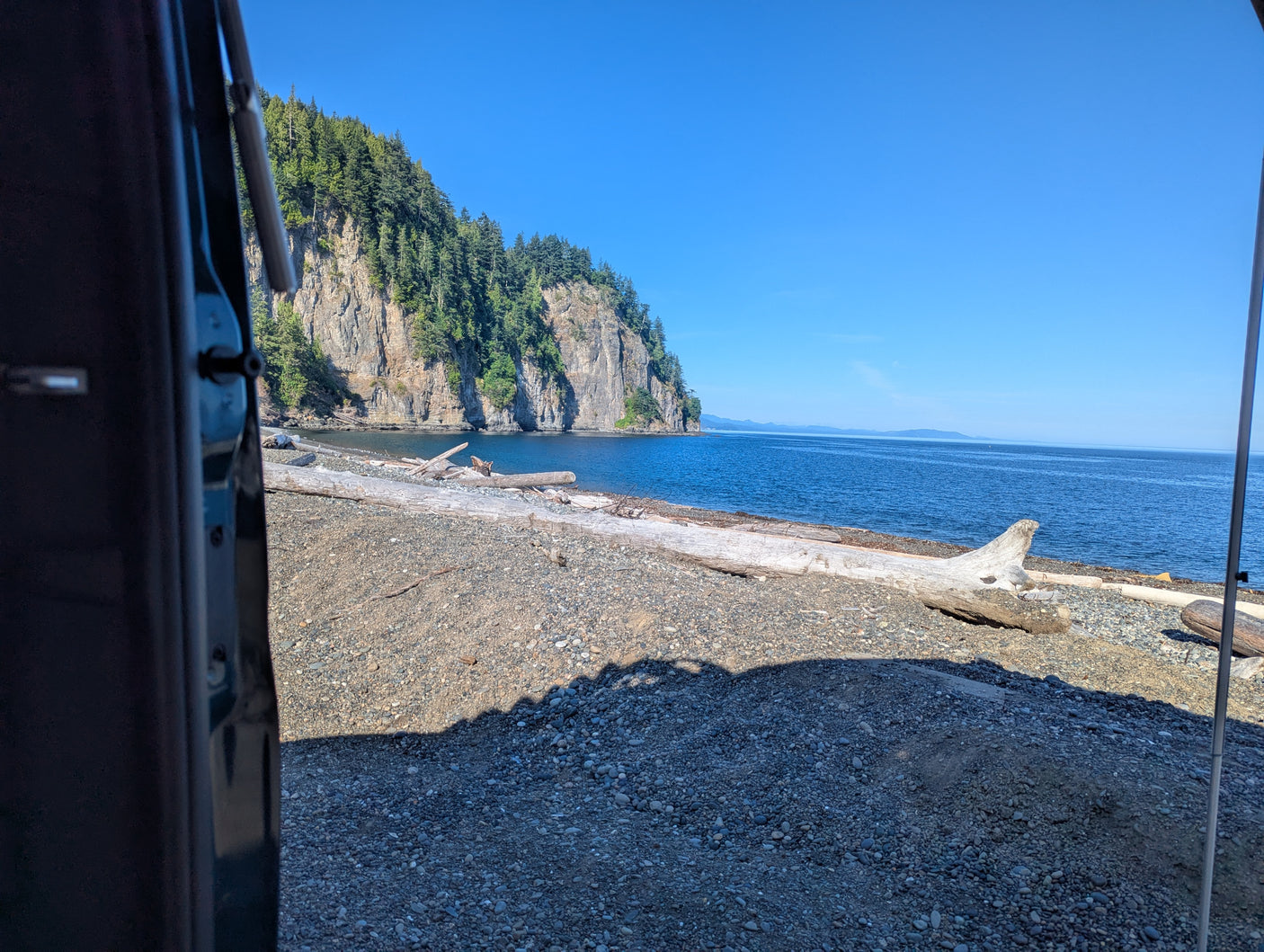 View from an RV rental: Rocky beach with driftwood, calm blue sea, and a tree-covered cliff.