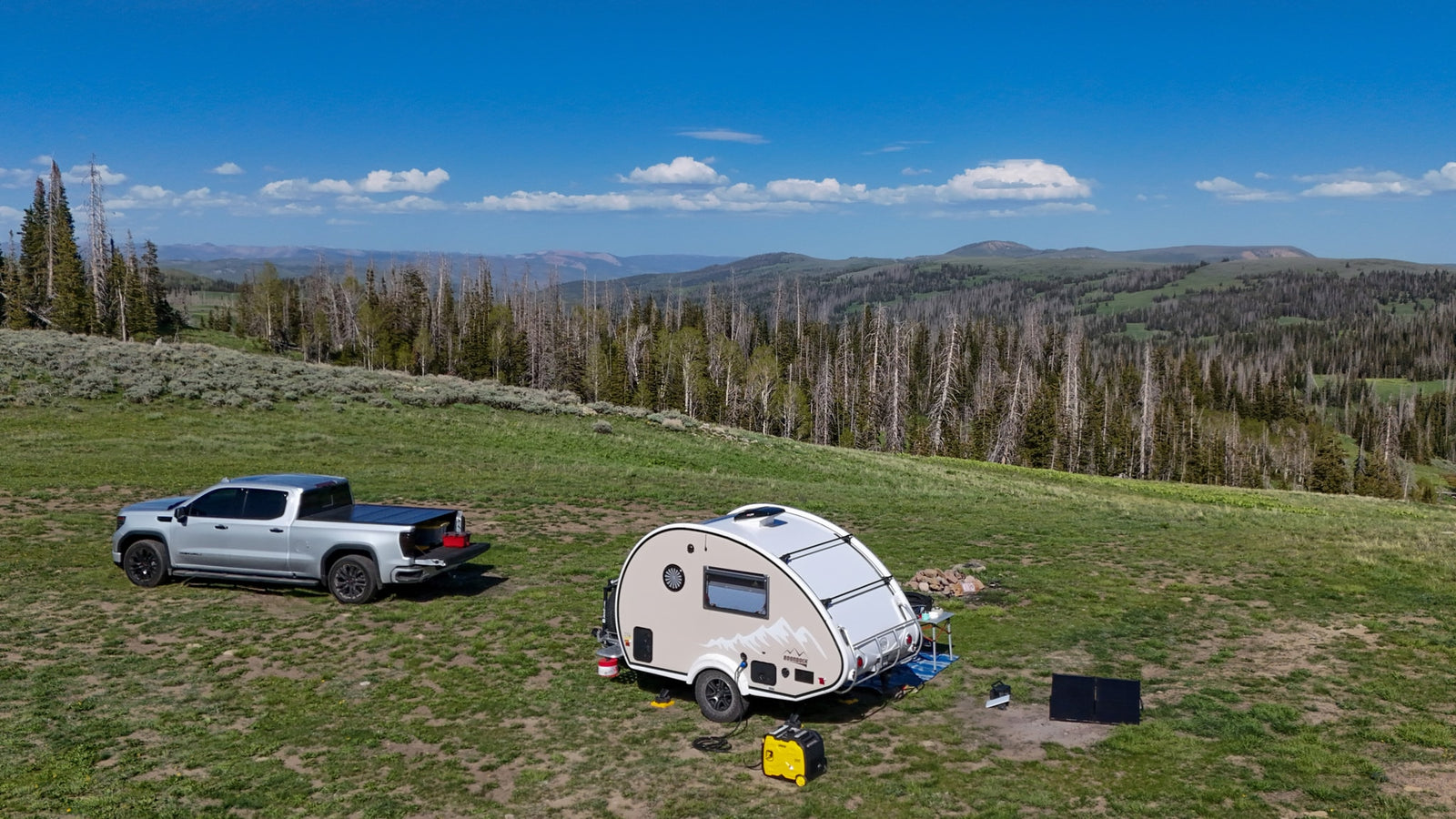 A silver pickup & teardrop camper are set up for off-grid RV rental camping on a grassy hill with distant mountains.