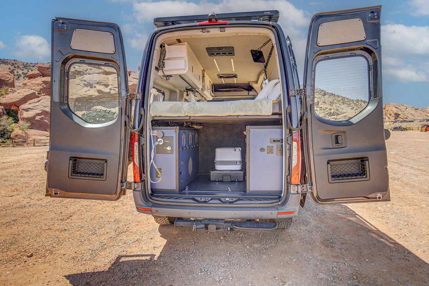 This rv rental camper van showcases its adventure-ready interior: a raised bed, organized storage, and portable toilet with open rear doors in a scenic, rocky desert.