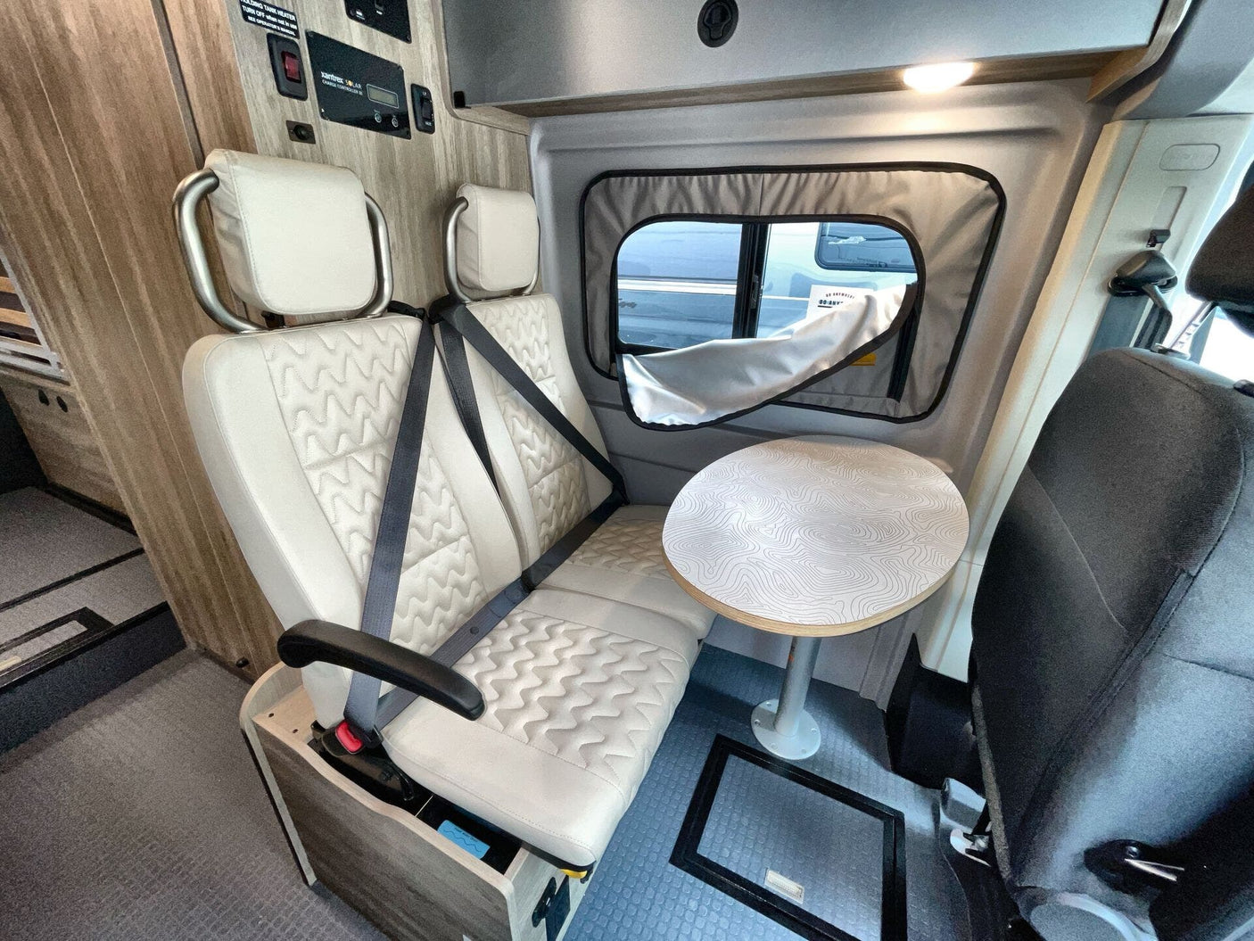 RV rental interior with cozy beige seats, a patterned round table, and a window. Controls for power system on the wall.