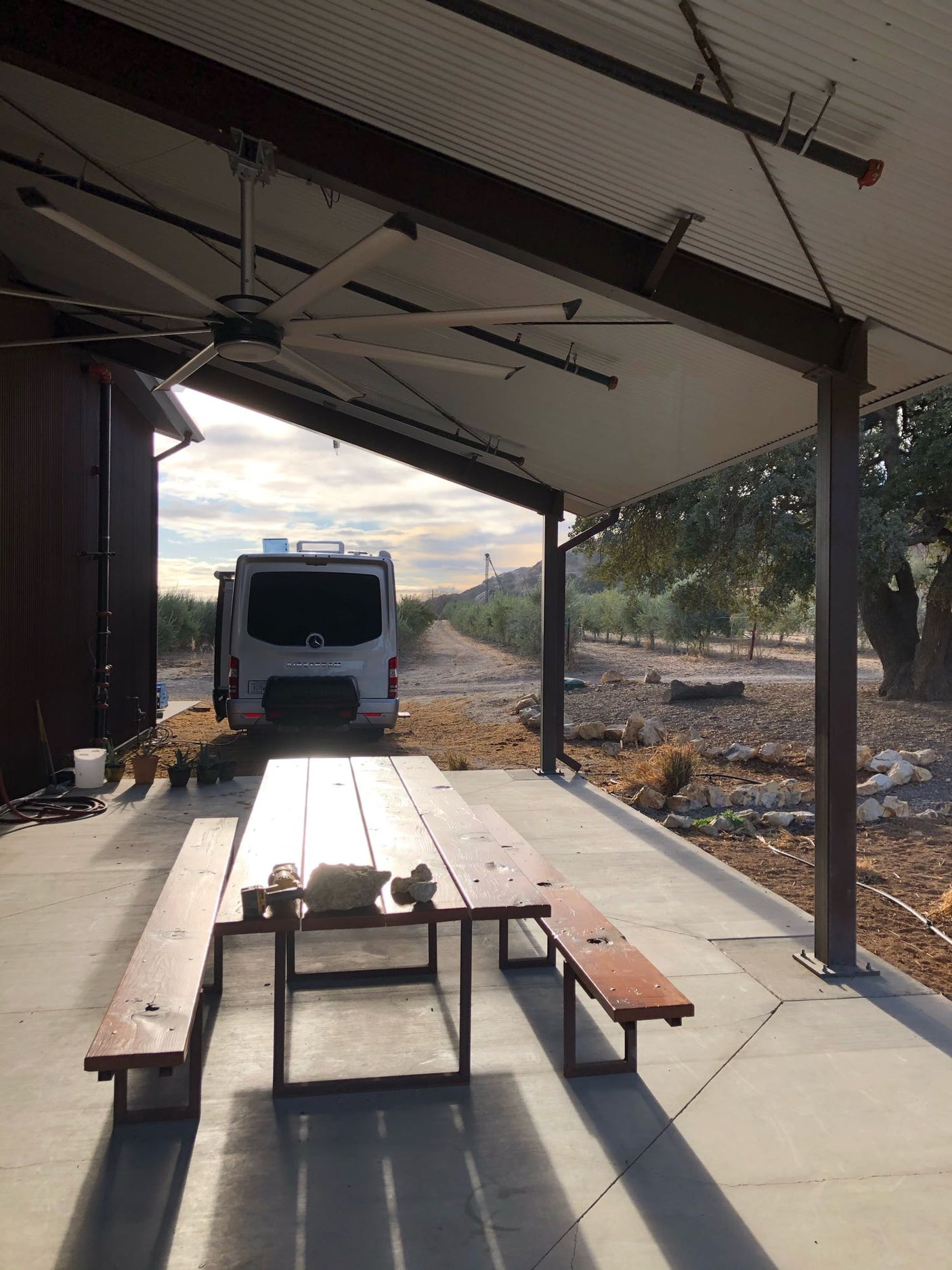 An RV sits by a building next to a shaded patio with a picnic table & large fan. Olive trees line the sunny rural landscape. Ideal for RV rental.