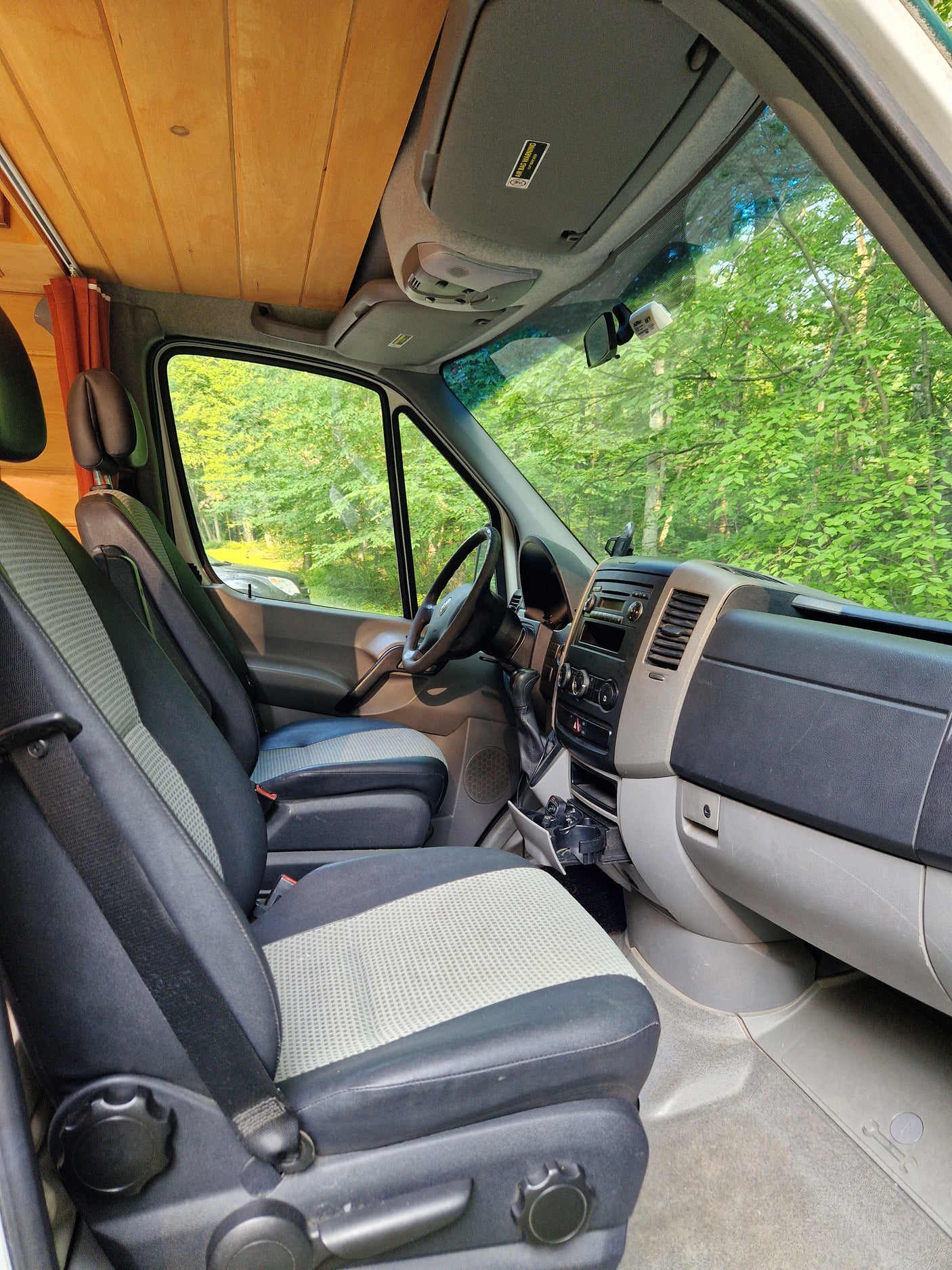 Front cabin of an RV rental: driver and passenger seats, dashboard, wooden ceiling, and green trees outside.