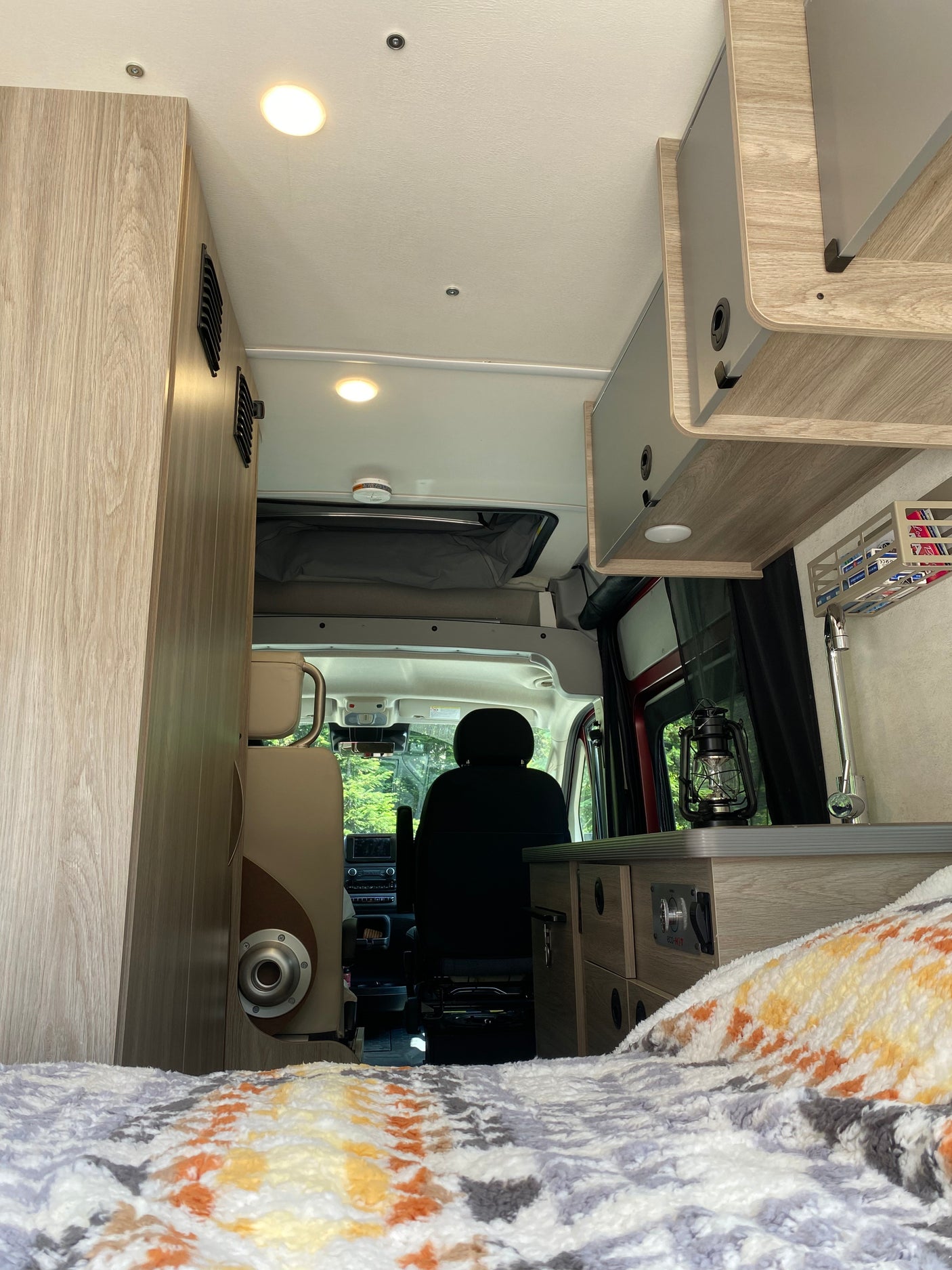 Cozy RV rental interior: light wood cabinetry, bed with colorful blanket, kitchen area, and driver's seat.