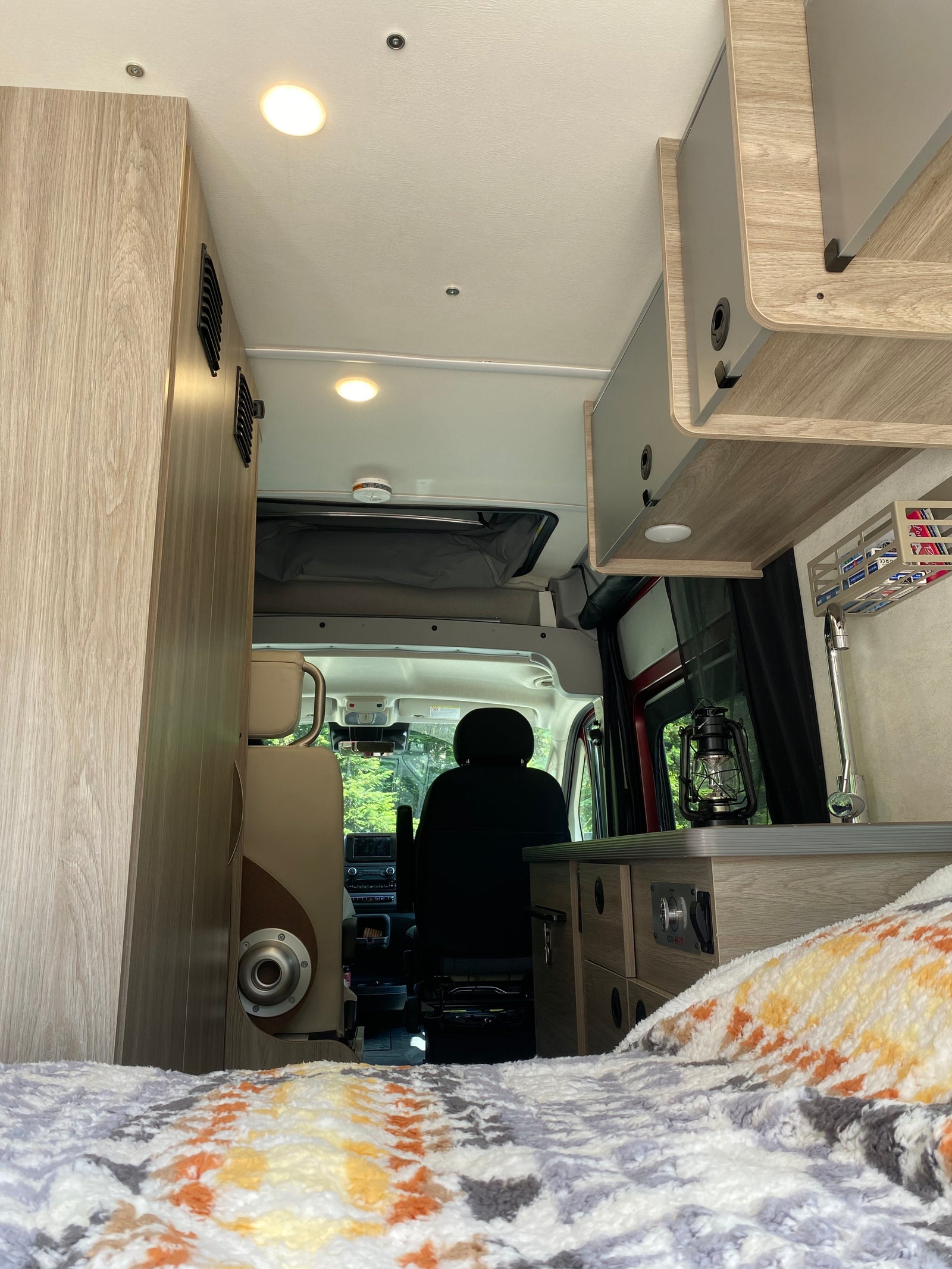 Cozy RV rental interior: light wood cabinetry, bed with colorful blanket, kitchen area, and driver's seat.