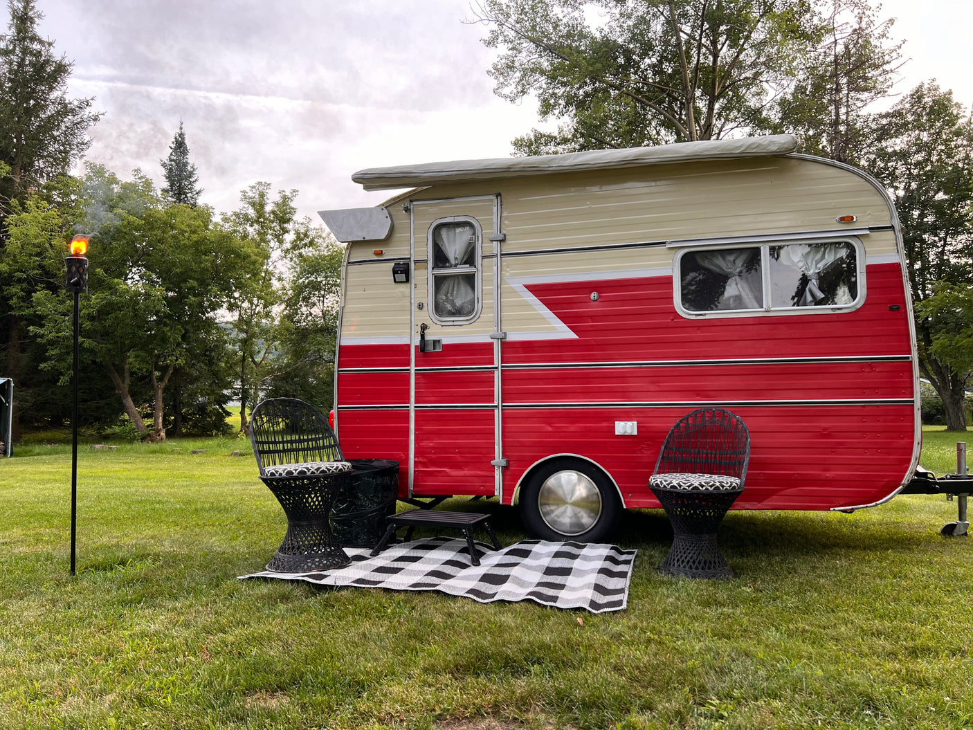 Cozy vintage RV rental with chairs, mat, and a lit tiki torch in a grassy field. Relax and enjoy the outdoors!