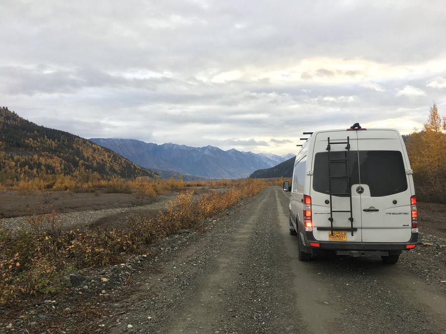Adventurous rv rental: a white Sprinter van on a gravel road in Alaska's autumn mountains.