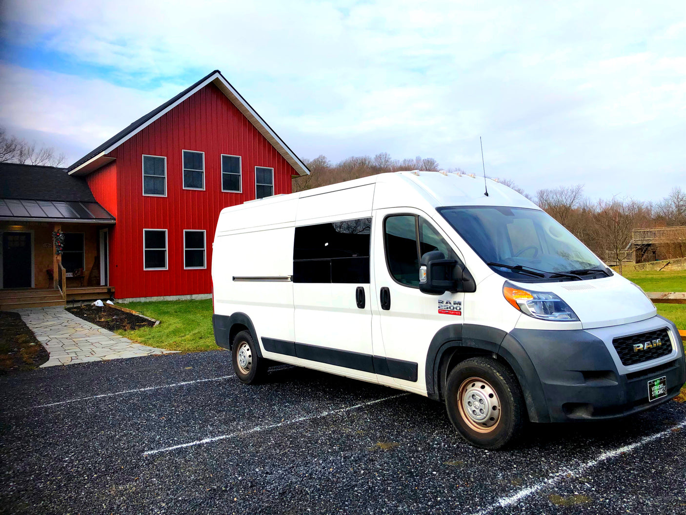 White Ram ProMaster camper van from our RV rental fleet, parked on a gravel lot beside a striking red building. Vermont views.