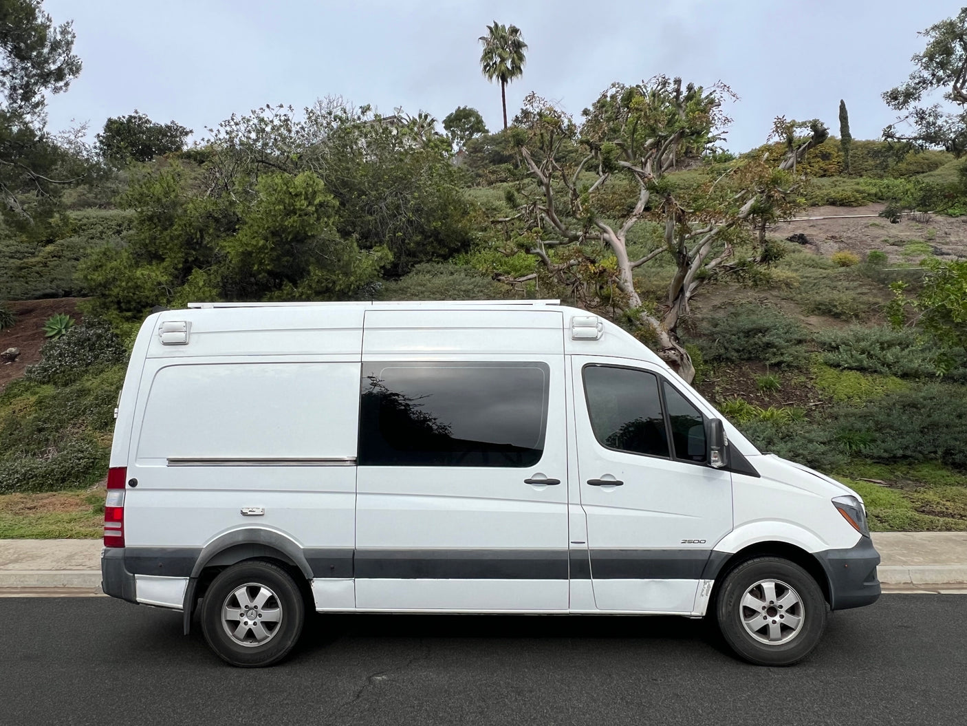 White Mercedes-Benz Sprinter camper van, great for an RV rental adventure, parked on a street with a lush green hillside.