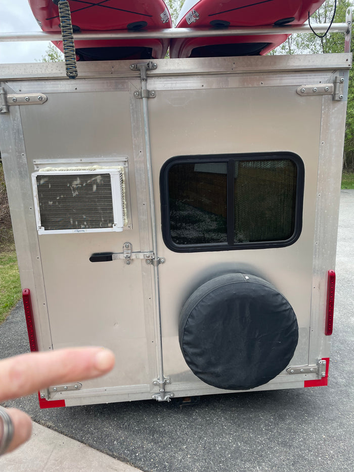 Rear view of a compact RV rental trailer, featuring kayaks overhead, a window AC unit, side window, and spare tire.