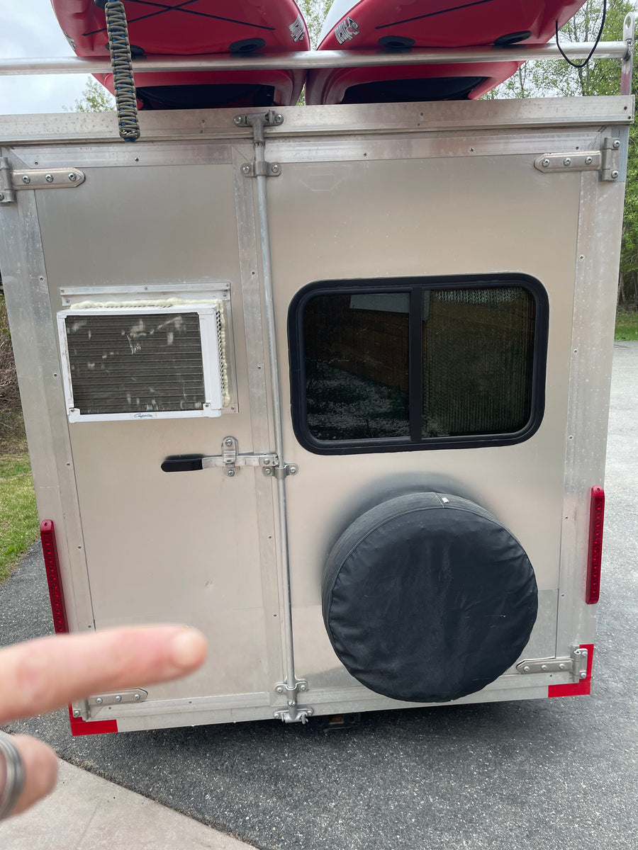 Rear view of a compact RV rental trailer, featuring kayaks overhead, a window AC unit, side window, and spare tire.