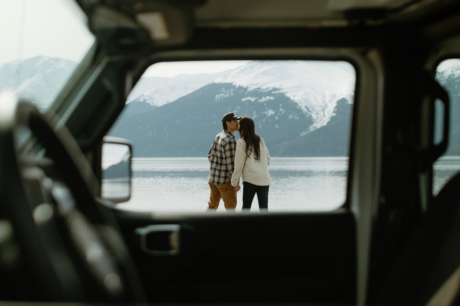 Inside an RV rental, a couple is framed by the back window, sharing a kiss by a scenic lake with mountains.