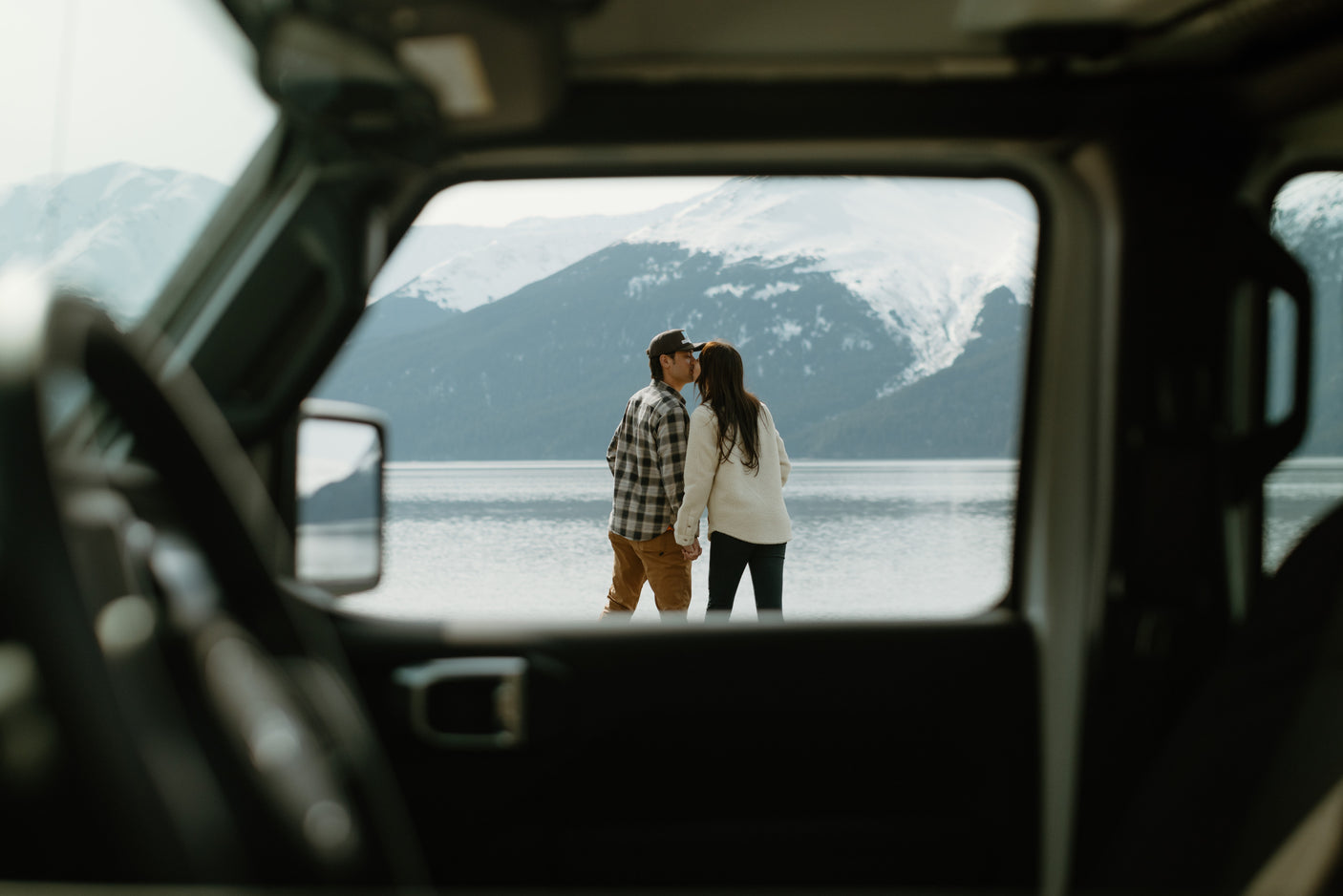 Inside an RV rental, a couple is framed by the back window, sharing a kiss by a scenic lake with mountains.