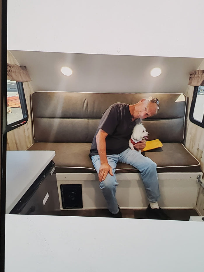 Inside an RV rental: A man gently holds his white dog while seated on a comfortable brown bench in the cozy camper.