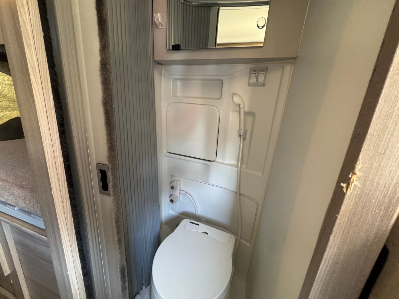 Compact RV rental bathroom featuring a white toilet, wall-mounted shower head, and small mirror above. — photo 7