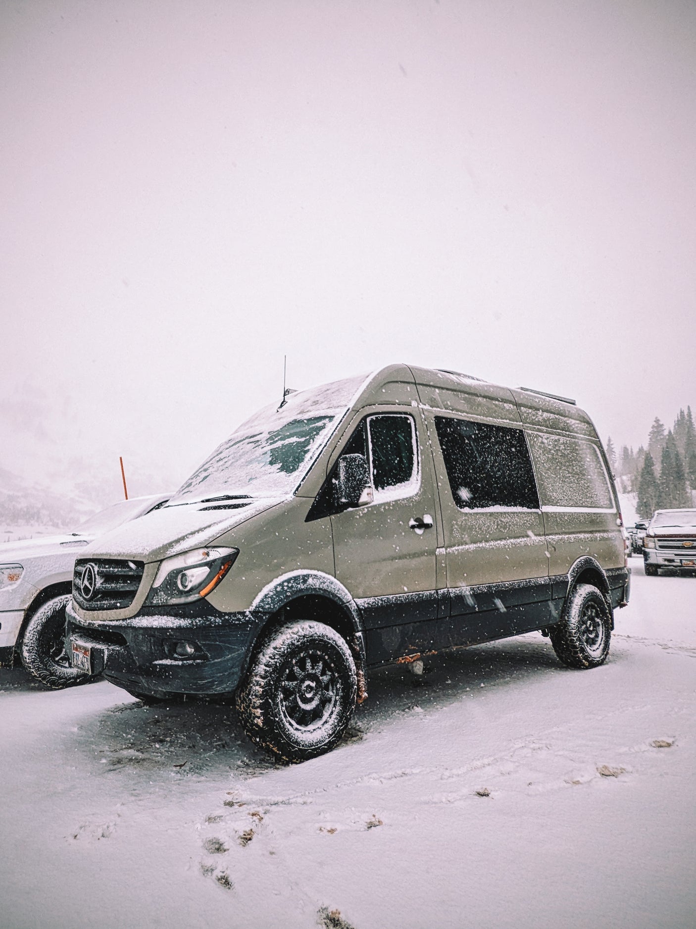 An olive green Mercedes Sprinter RV rental, fitted with large off-road tires, stands ready in a snowy winter landscape.