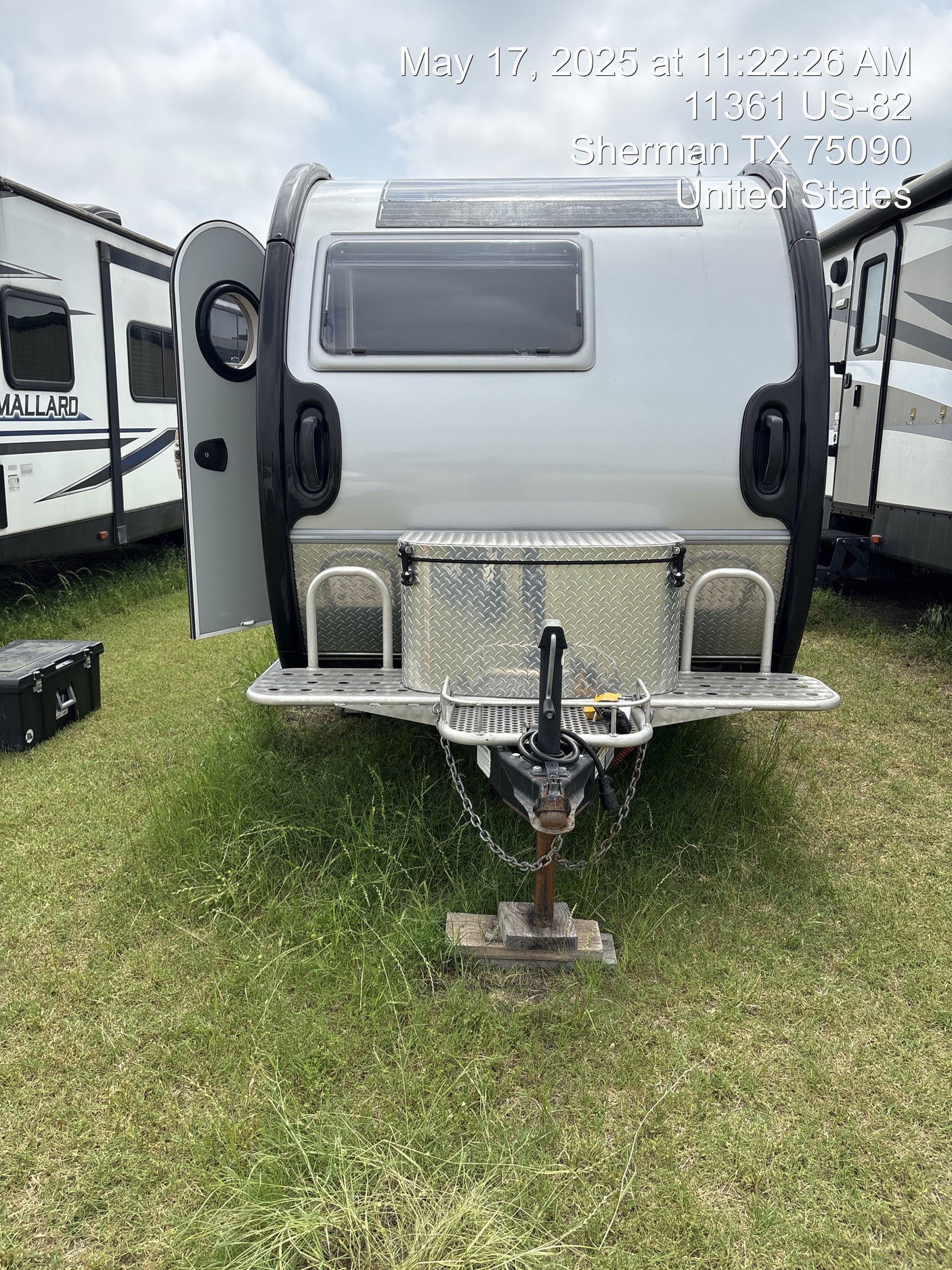 A sleek silver travel trailer, ready for RV rental, is parked on a grassy lot with other camping vehicles nearby.