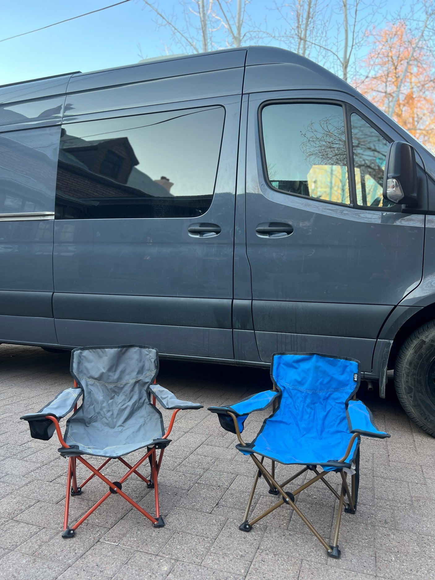 A grey rv rental van with two folding camping chairs. The van's windows reflect a house and trees, suggesting a travel setup.