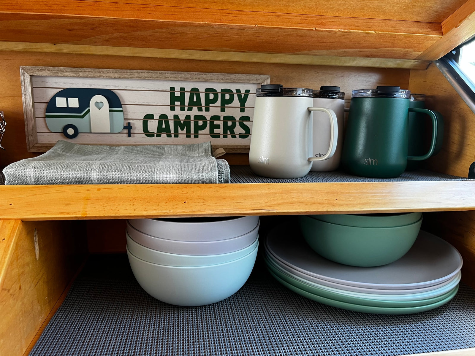 Organized RV rental kitchen with "HAPPY CAMPERS" decor, travel mugs, and stacked dishware neatly stored on shelves.