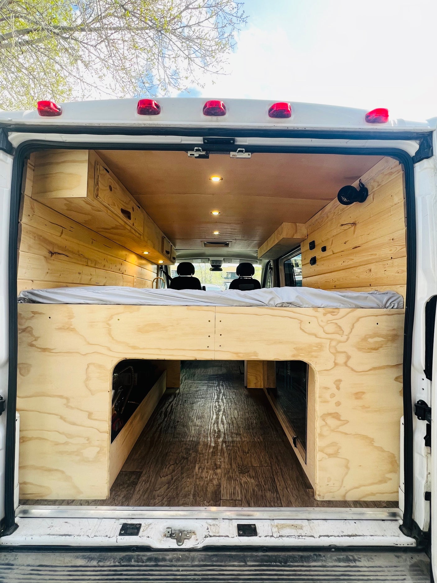 View of an RV rental van's interior: a wooden bed platform with grey mattress, under-bed storage, wood paneling, and ceiling lights.