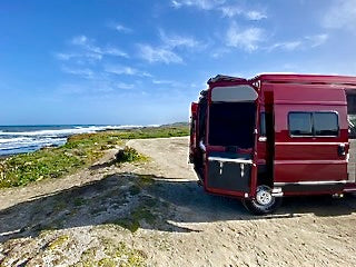 Imagine your next adventure: a red RV rental parked by the ocean under a blue sky, its side door open to the breeze.
