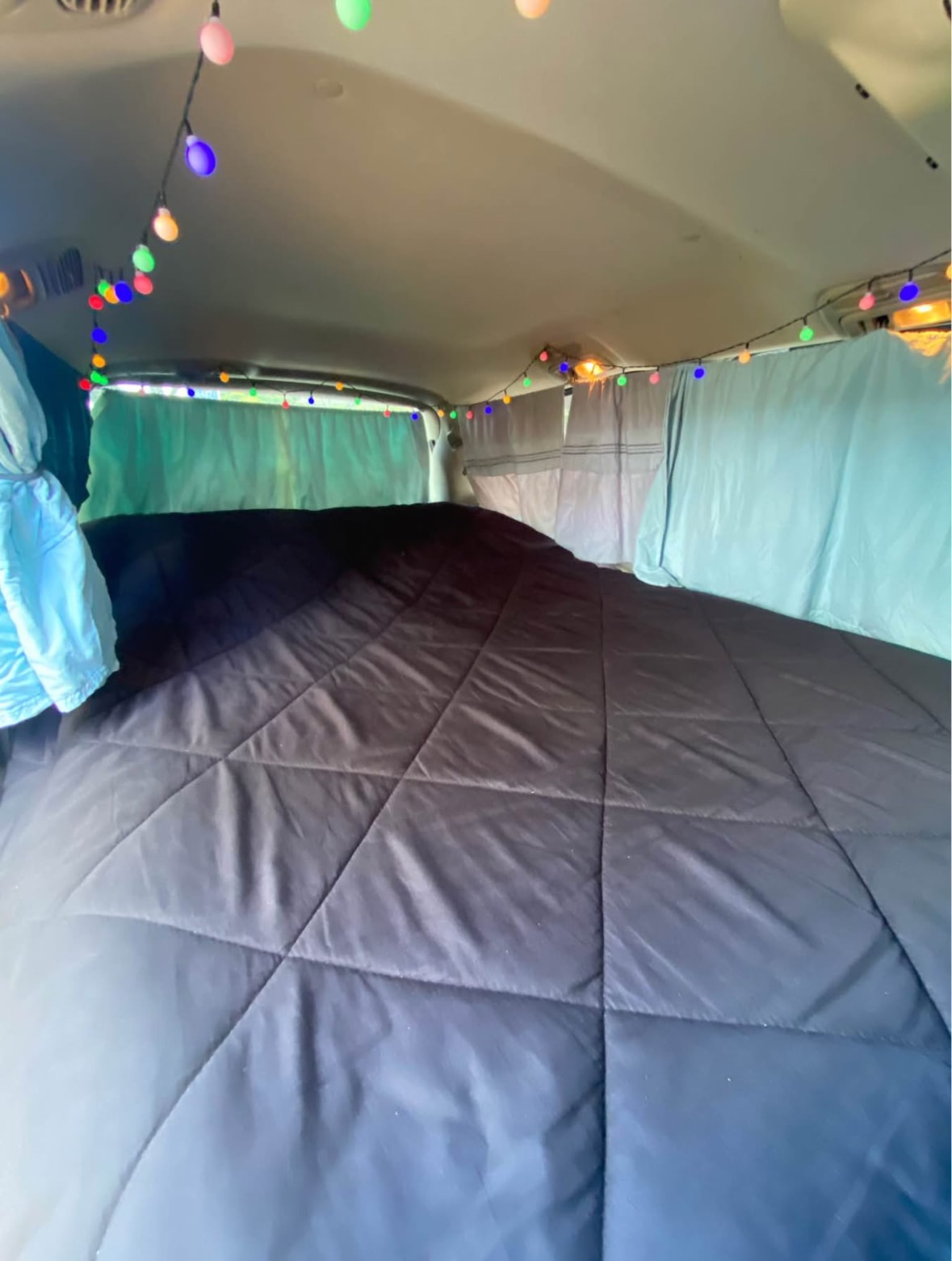 Cozy RV rental interior with a dark quilted bed, blue curtains, and colorful string lights for a comfortable road trip.