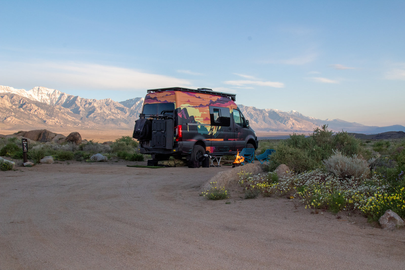 This vibrant RV rental is parked at a desert campsite, ready for adventure. Campfire glowing with stunning snow-capped mountains behind.