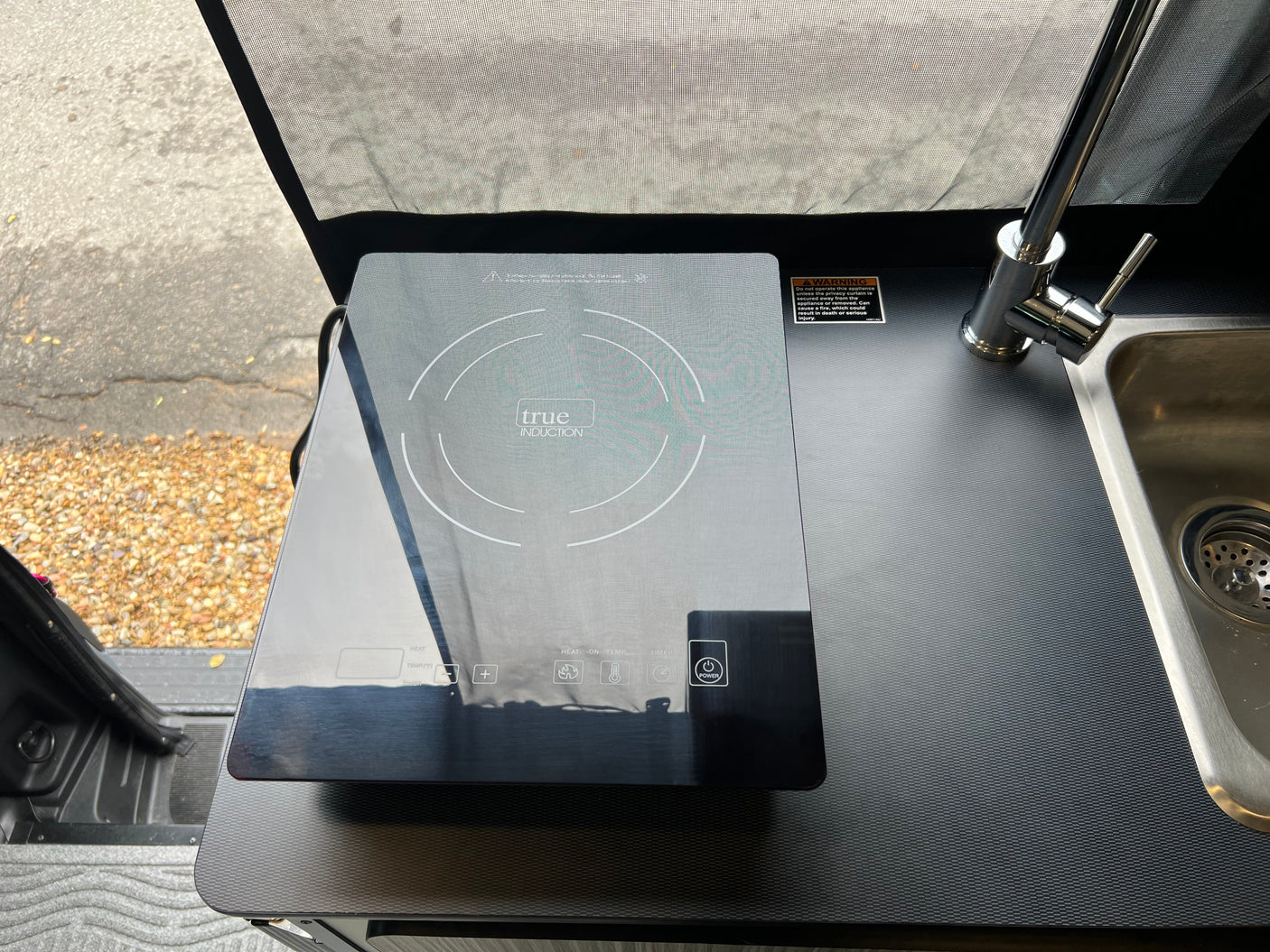 RV rental kitchenette with a sleek True Induction cooktop and sink. Includes safety warning about a nearby curtain.