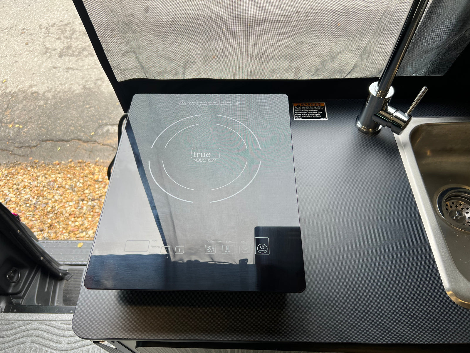RV rental kitchenette with a sleek True Induction cooktop and sink. Includes safety warning about a nearby curtain.