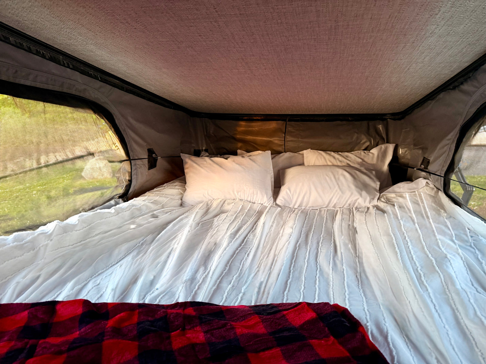 A comfy bed with white bedding & a red plaid blanket inside an RV rental tent, offering views of green outdoors.