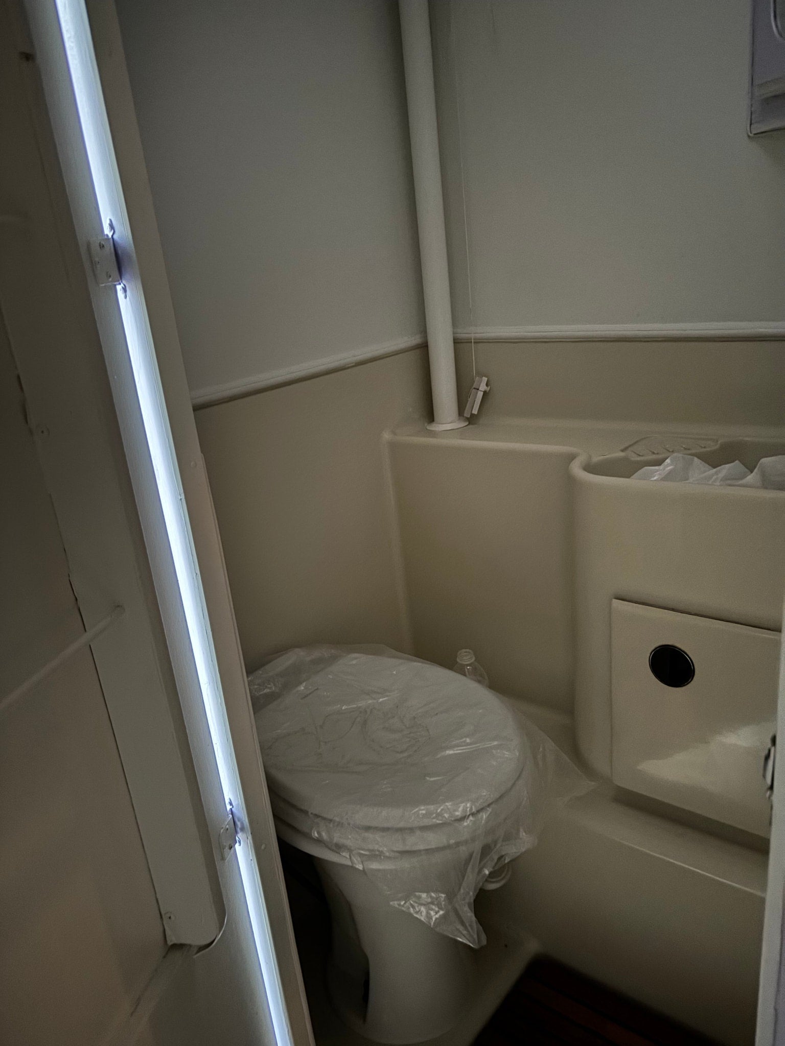 Compact RV rental bathroom with a new toilet (covered in plastic), integrated sink/shower, and bright vertical lighting.