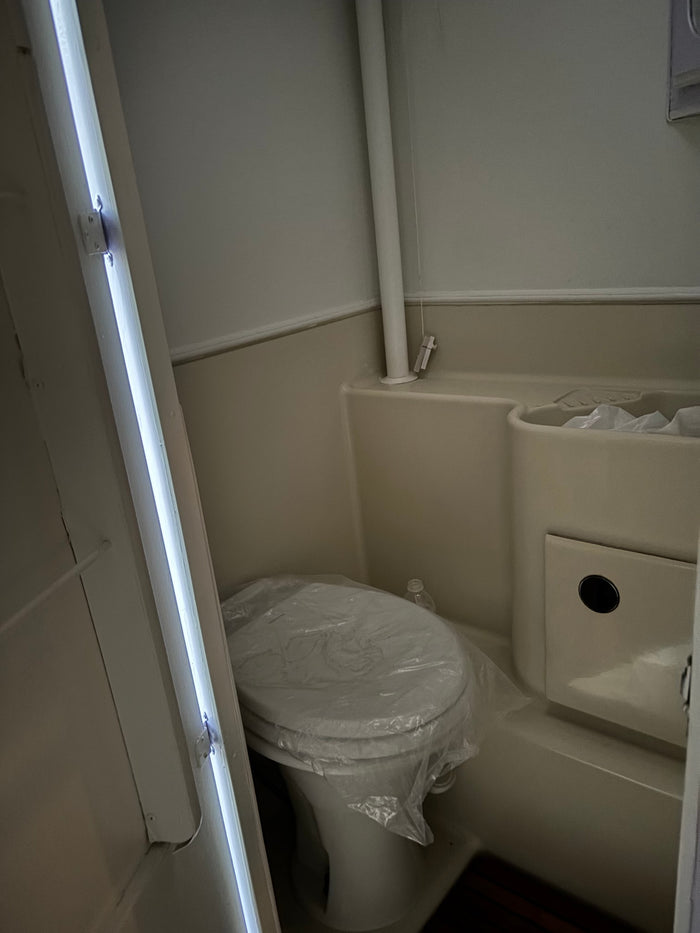 Compact RV rental bathroom with a new toilet (covered in plastic), integrated sink/shower, and bright vertical lighting.