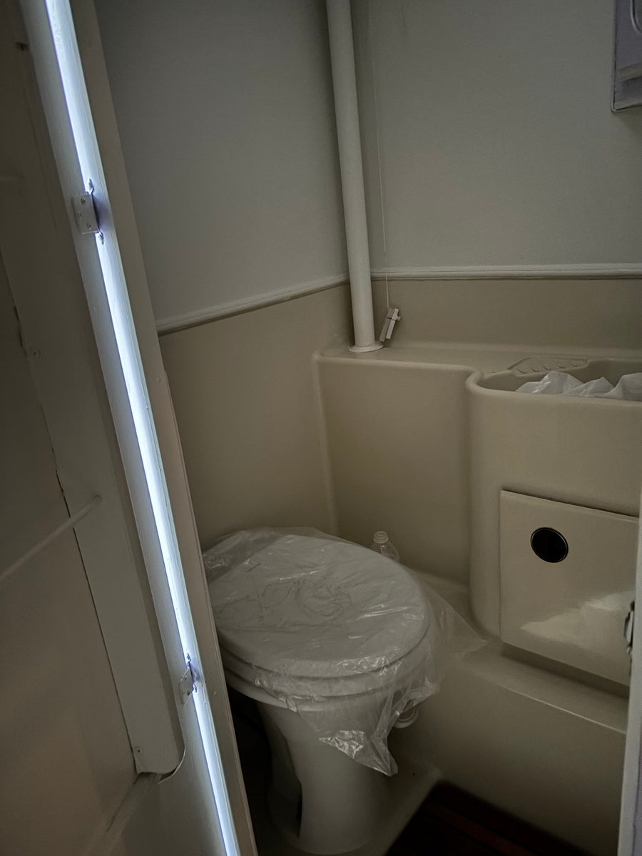 Compact RV rental bathroom with a new toilet (covered in plastic), integrated sink/shower, and bright vertical lighting.