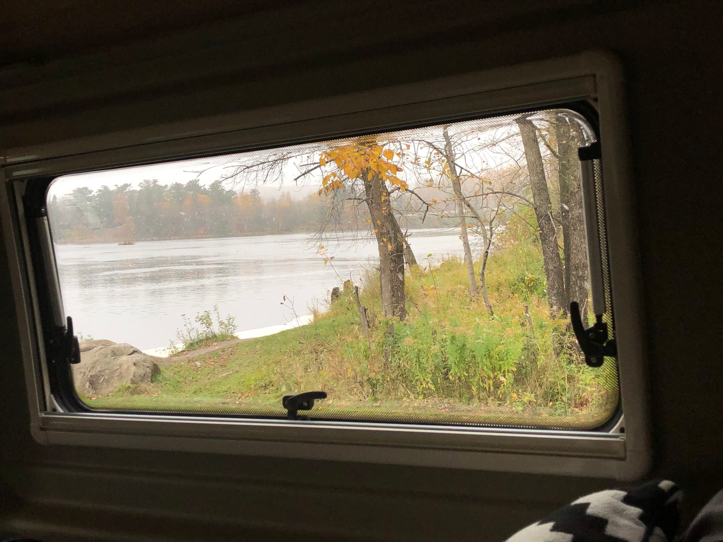 Scenic RV rental window view of a quiet river, autumn foliage, and falling snow on a cloudy day.
