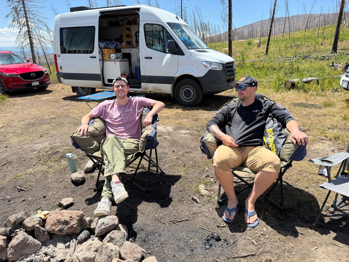 Two men relaxing at a campsite with an RV rental and a red SUV, near a fire pit in a scenic, treed area. — photo 5