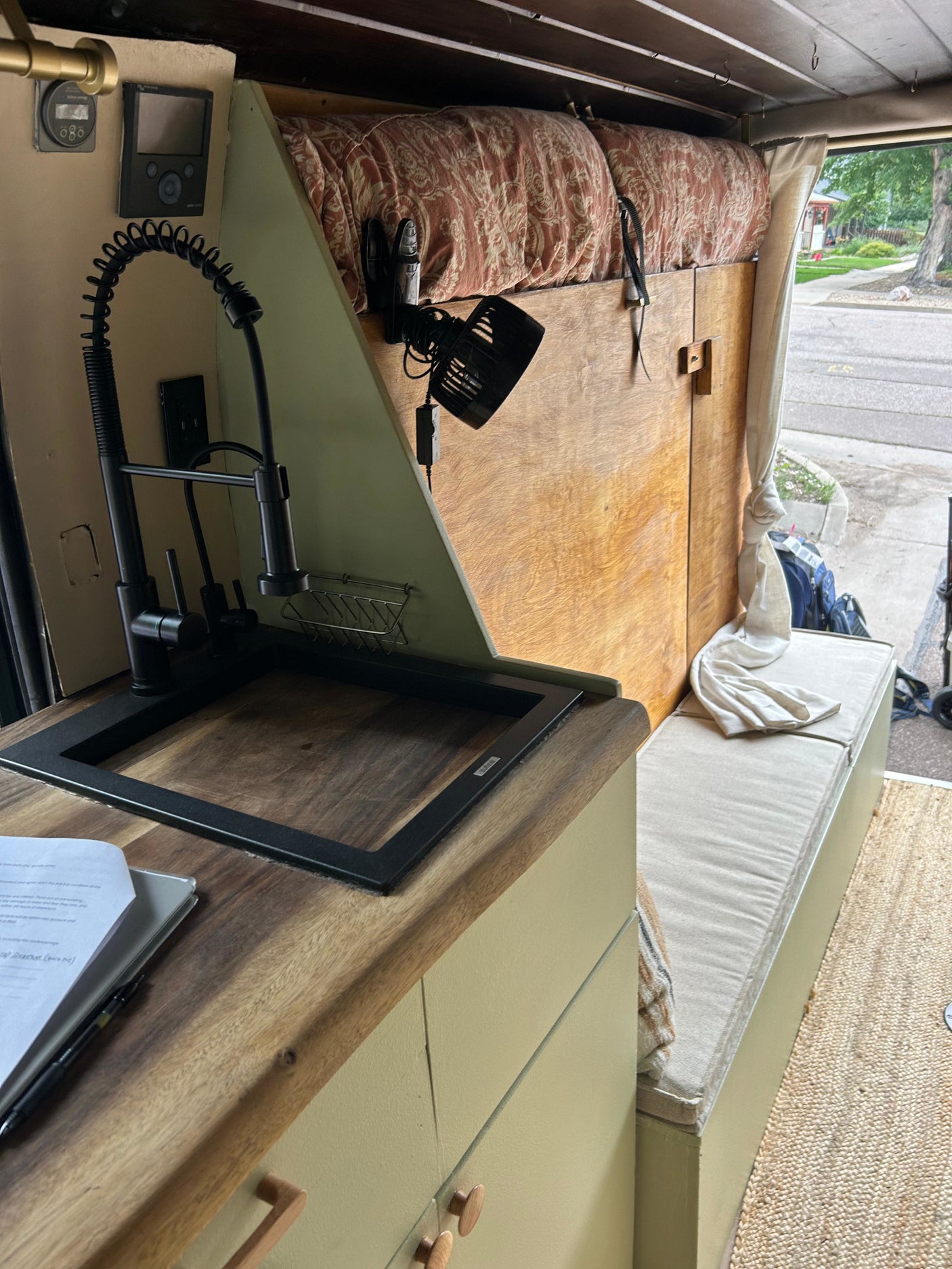 Inside a cozy rv rental: kitchen counter, sink, controls, and bed with patterned cushion. View of street outside.