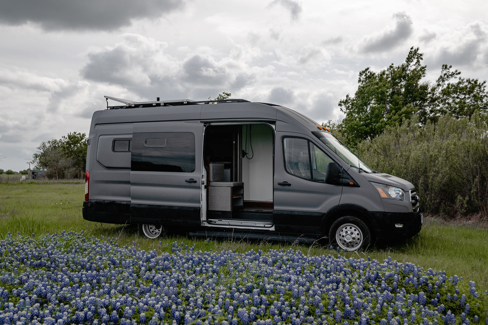 A grey camper van (RV rental) with its side door open is parked amidst a field of blue flowers under a cloudy sky.