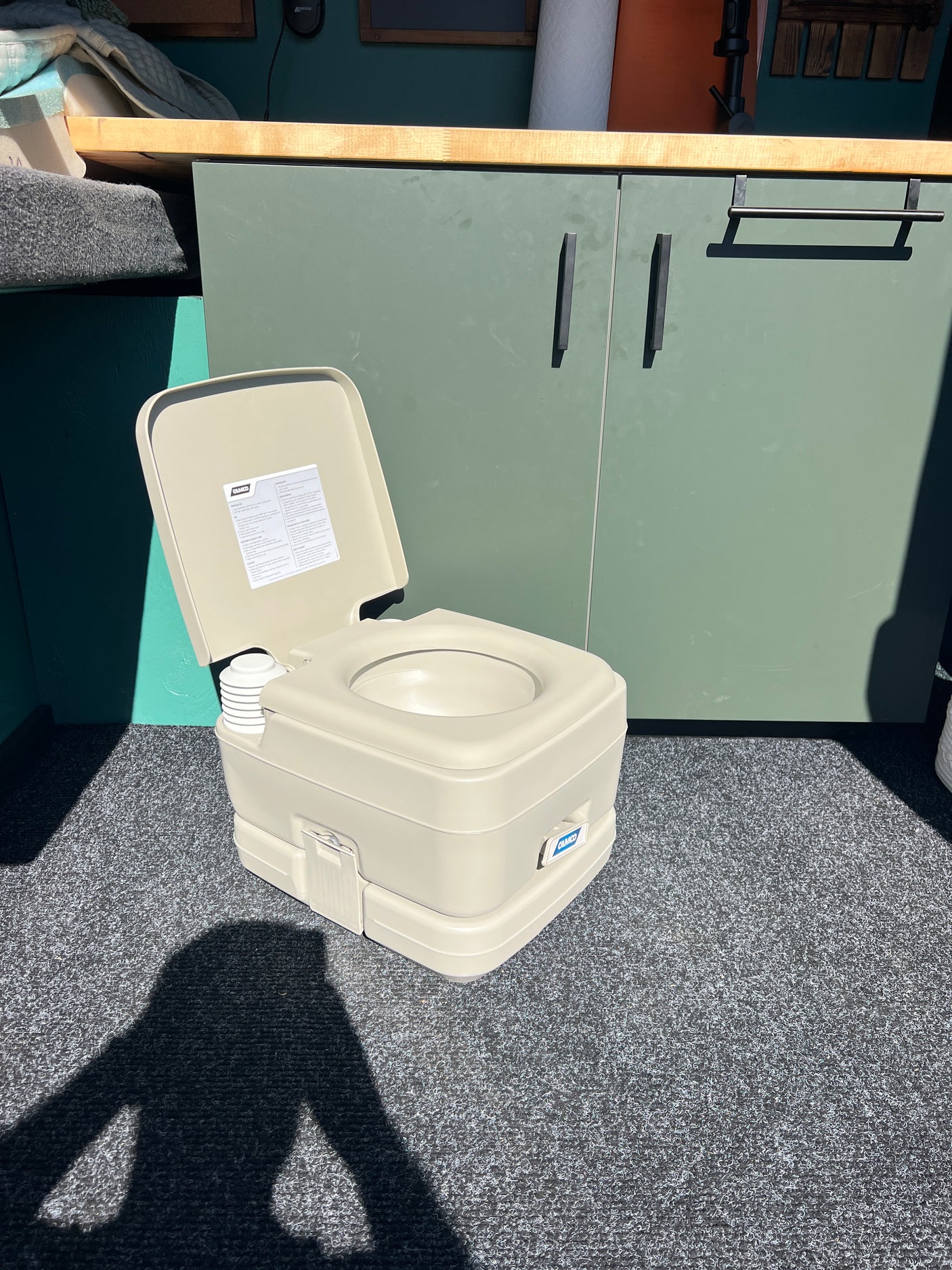 RV rental amenity: A portable CAMCO toilet with lid open, on gray textured flooring, inside a camper's green cabinet space.