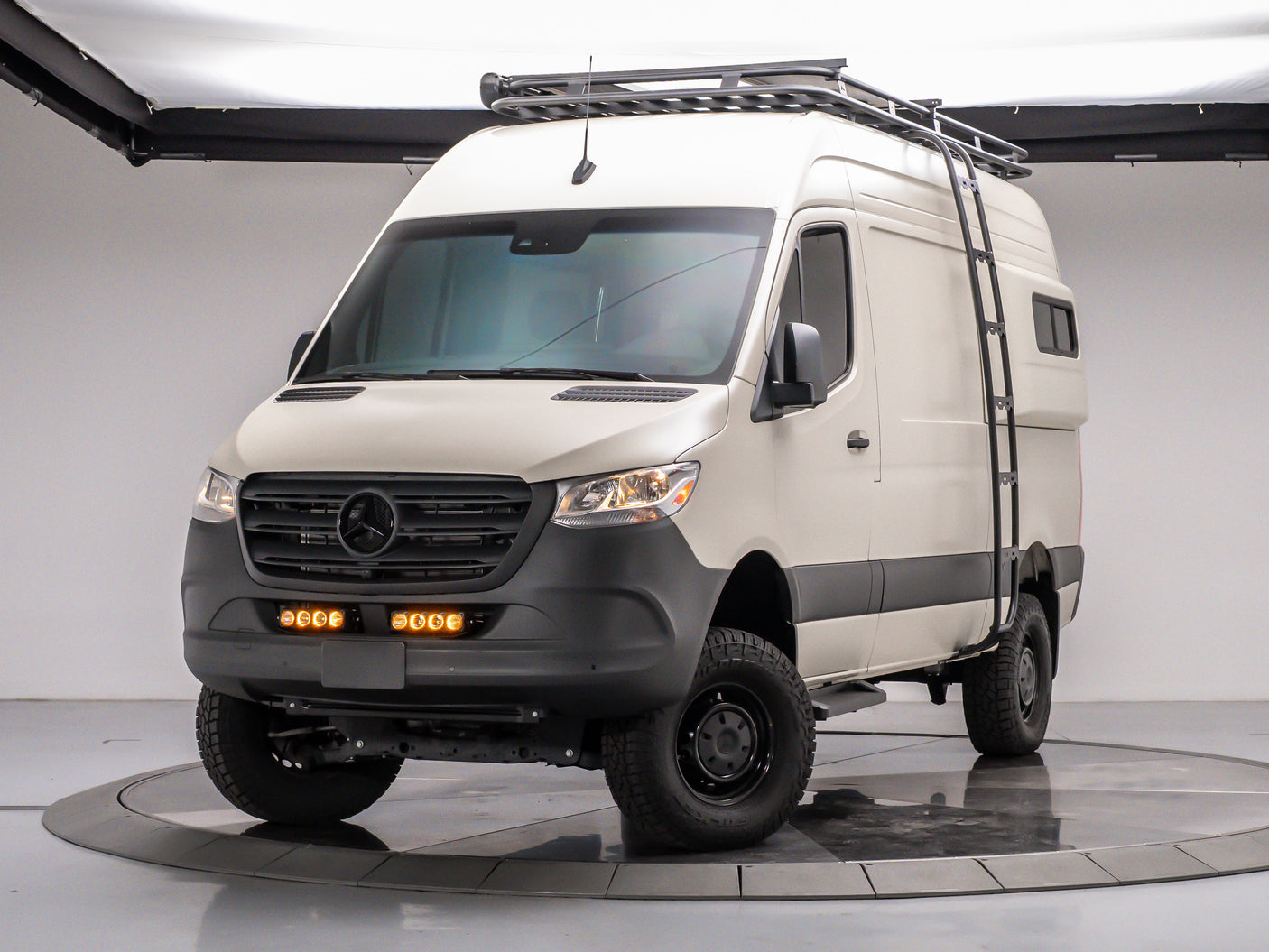 White Mercedes Sprinter adventure van, equipped with off-road tires, roof rack, ladder, and amber lights. Ideal for **rv rental**.