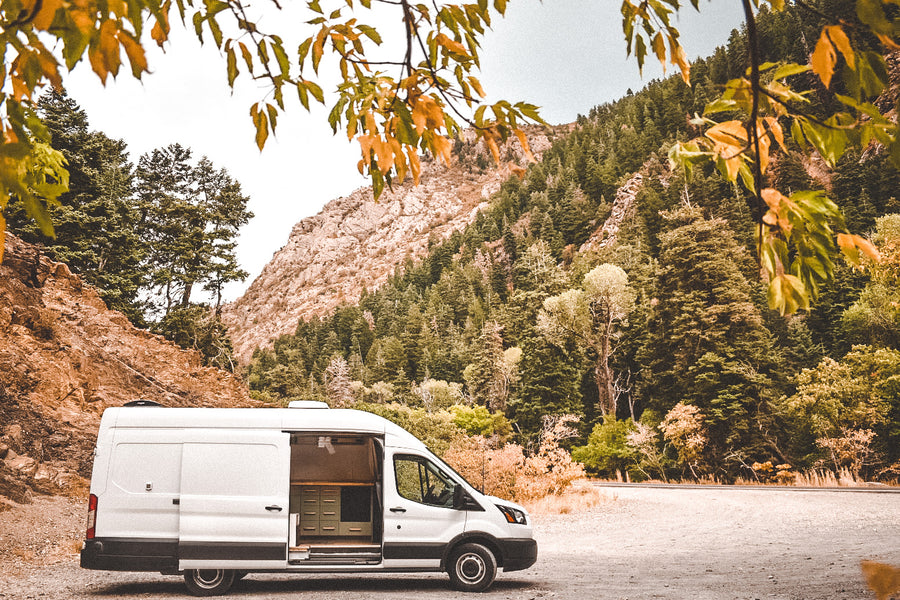 A white camper van, available for RV rental, parked on a dirt road amidst stunning mountain forests.