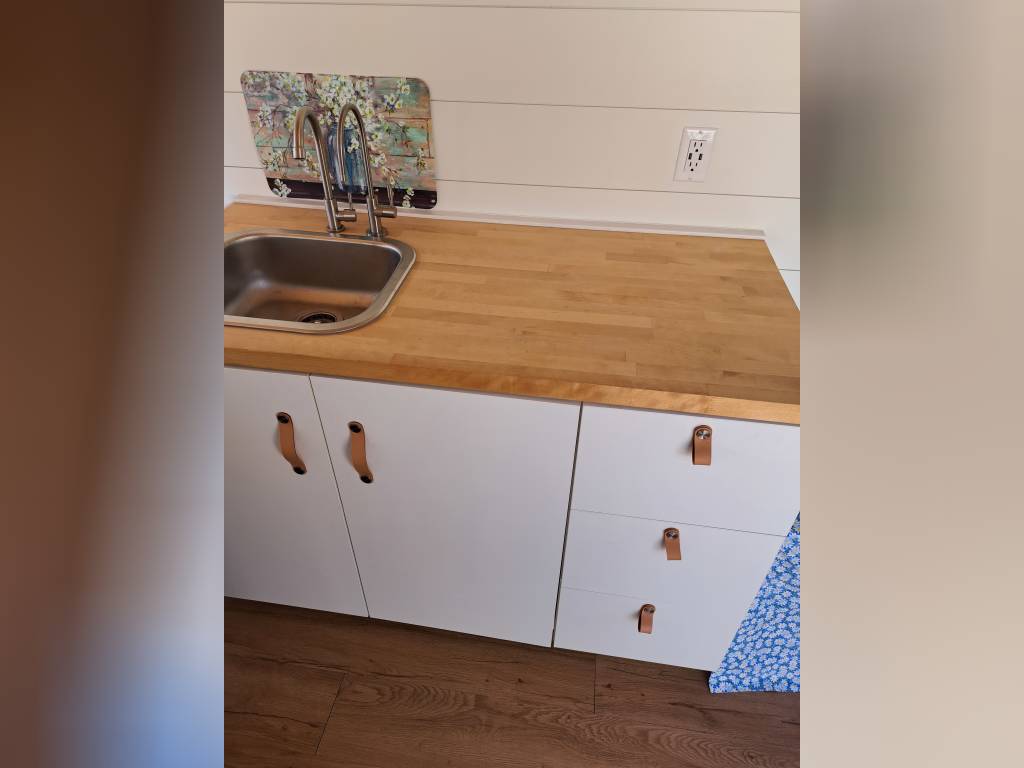 and compact fridge.|Open side of a white camper van shows its custom interior with kitchen and seating