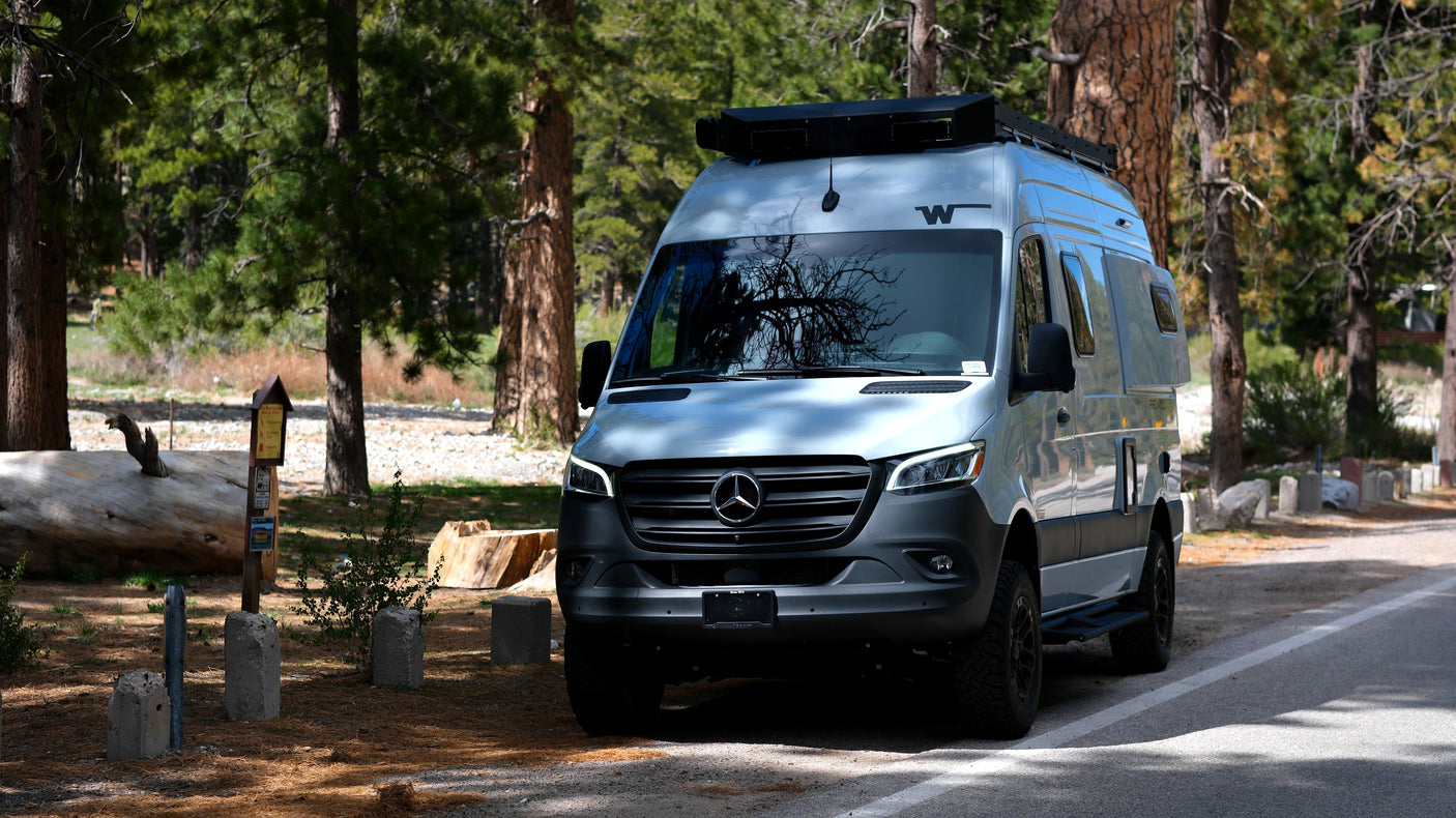 Light blue Winnebago Mercedes-Benz Sprinter RV rental camper van with roof rack, parked on a paved road in a pine forest.